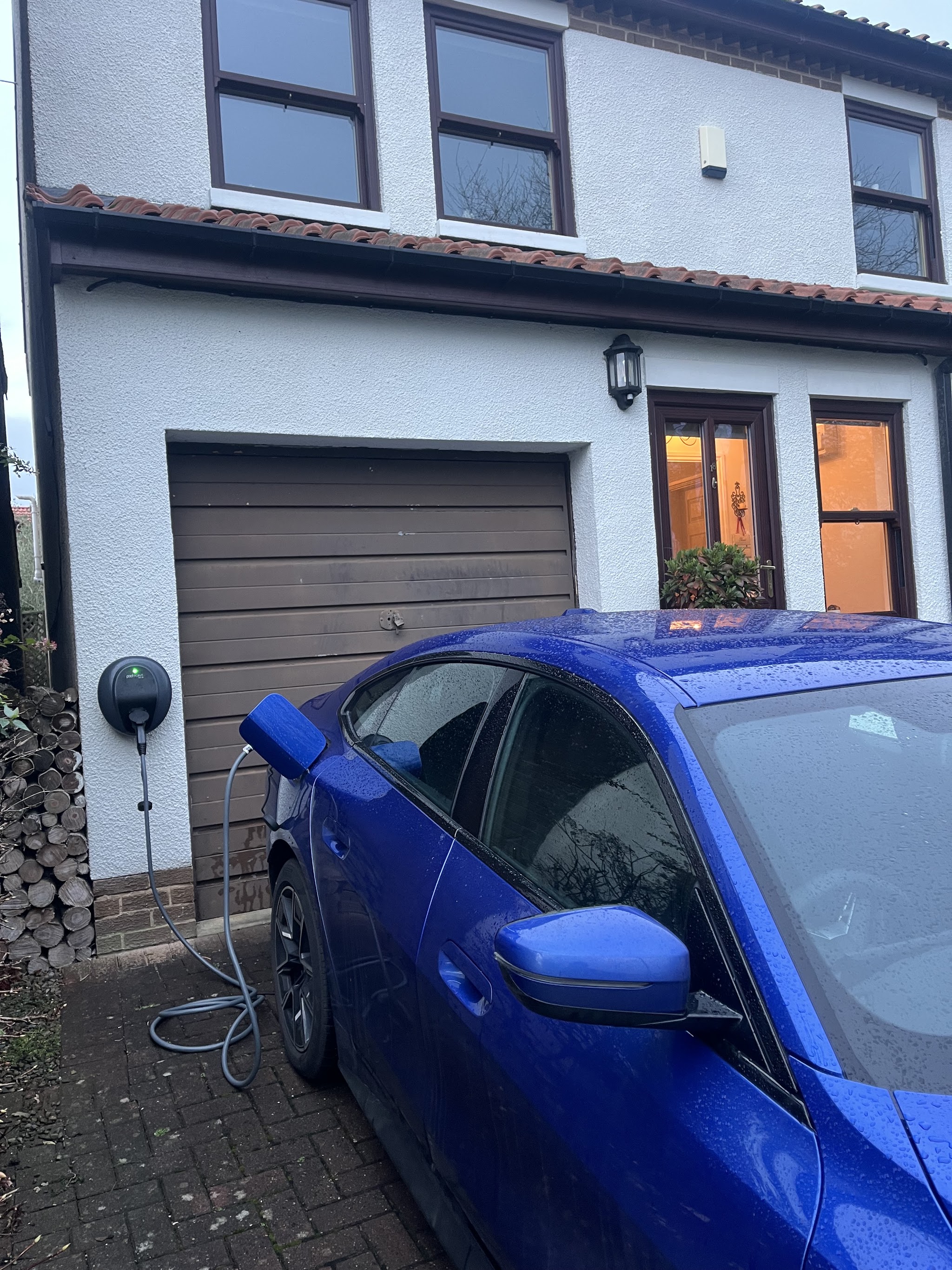 Home EV charger on the front wall of a house, plugged into a blue car on the driveway